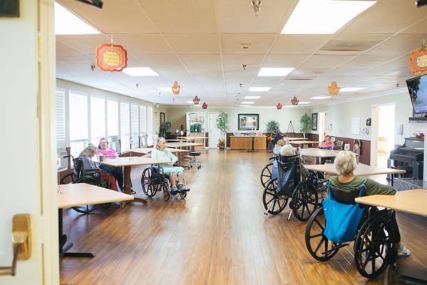Residents in wheelchairs in a spacious common area with tables.
