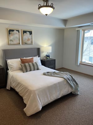 A cozy, well-lit bedroom featuring a queen-sized bed, two decorative pillows, and a window with natural light.