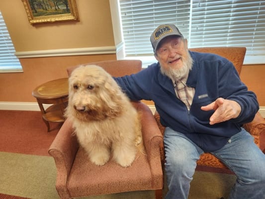 An elderly man sitting next to a fluffy dog on a chair