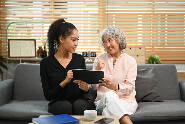 A senior woman and a caregiver interact over a tablet in a cozy living room.