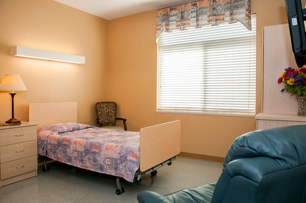 A bedroom in a senior living facility featuring a bed, chair, lamp, and dresser.