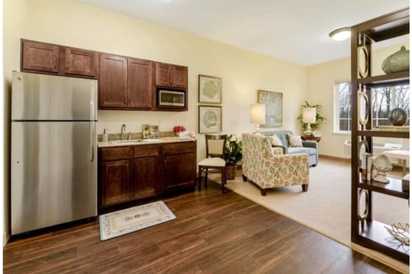 A kitchen and living area in a senior living facility featuring a refrigerator, cabinets, and a comfortable seating area.