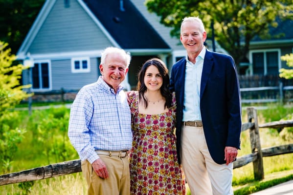 Three people standing outside a facility with a house in the background