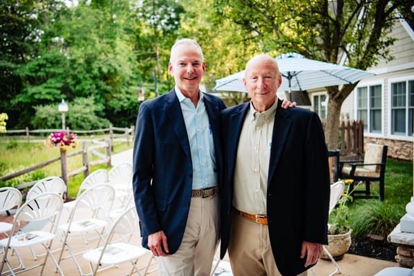 Two men pose together outside in a garden setting.