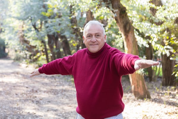 A smiling elderly man with arms extended in a scenic outdoor setting