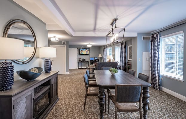 Bright dining area with a large table and chairs, featuring lamps and a television.