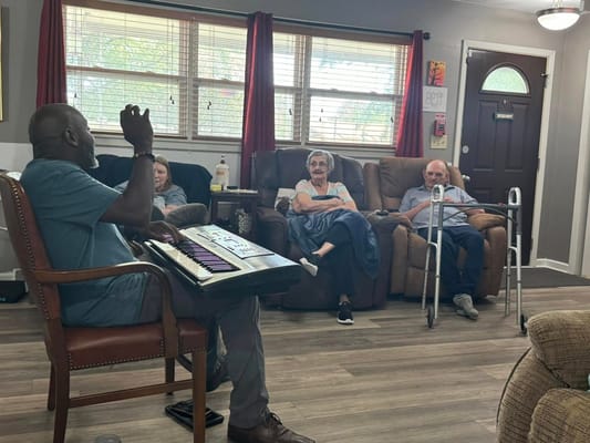 A musician plays a keyboard while seniors enjoy the performance in a cozy living room.
