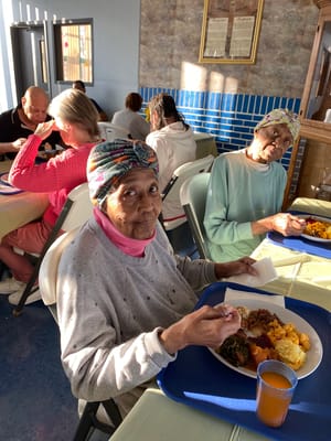 Two elderly women dining at a communal table with food in front of them