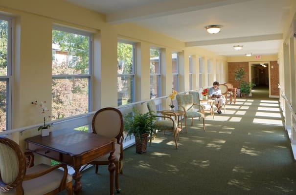 A well-lit hallway with chairs and tables, featuring windows and greenery.