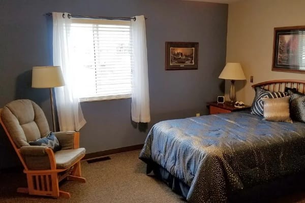 A cozy bedroom with a bed, armchair, and window with blinds