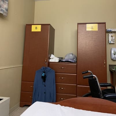 A room containing two wooden wardrobes labeled A and B, a wheelchair, and a blue coat.