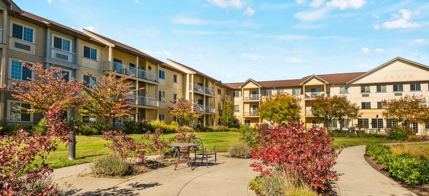 A sunny courtyard at Crescent Park Senior Living with blooming plants and two buildings in the background.