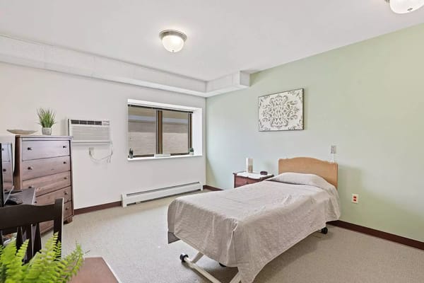 A well-furnished bedroom with a bed, dresser, and window at Sun Prairie Senior Living.