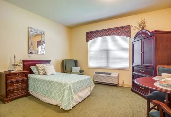 Well-furnished bedroom with a bed, chair, and table in a senior living facility.