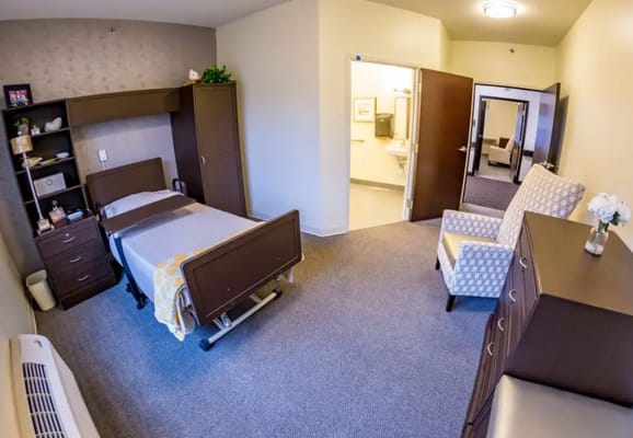 A spacious bedroom in a senior living facility featuring a bed, armchair, and wardrobe.