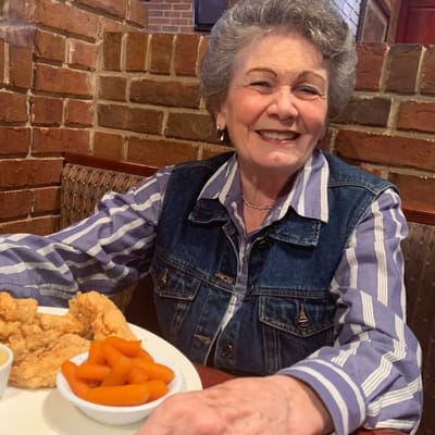 A smiling elderly woman enjoying a meal with fried chicken and carrots