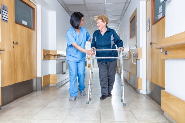 A nurse assisting an elderly woman with a walker in a hallway