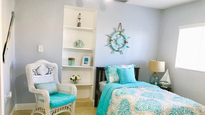 A cozy bedroom with a blue and white theme, featuring a bed, a chair, and decorative shelves.