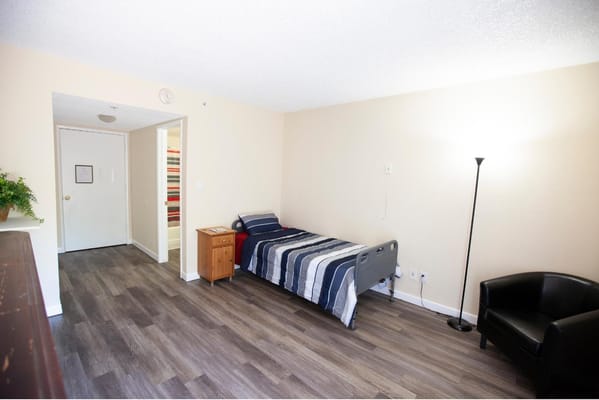 A room with a bed, nightstand, and chair in Rose Court Senior Living