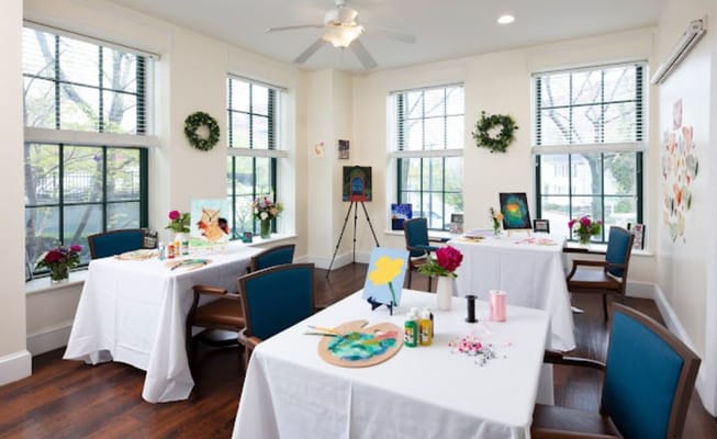Bright art studio with tables and flowers, featuring paintings and art supplies.
