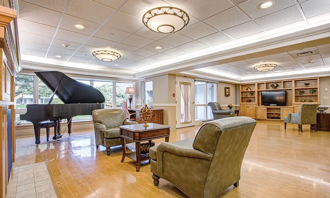 Cozy lounge with a piano, chairs, and a TV in a senior living facility.