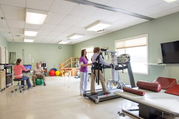 A senior assistant on a treadmill with guidance from a staff member in a therapy room.