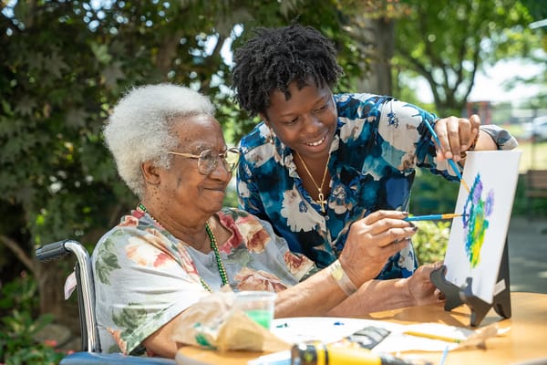 An elder woman painting outdoors with staff assistance