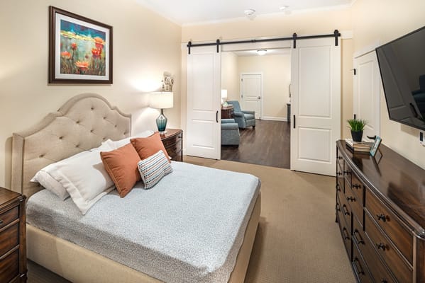 A cozy bedroom featuring a bed with decorative pillows, a dresser, and a sliding door to the living area.