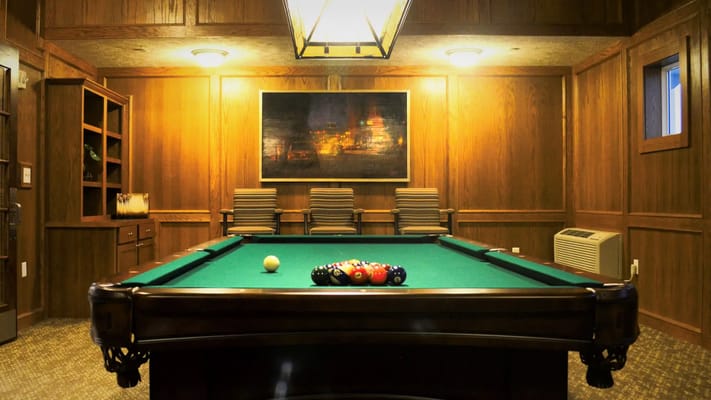 A well-lit pool table room with green felt, balls arranged for play, and wooden paneling.
