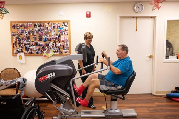 Senior man using an exercise machine with staff assistance in a communal area.