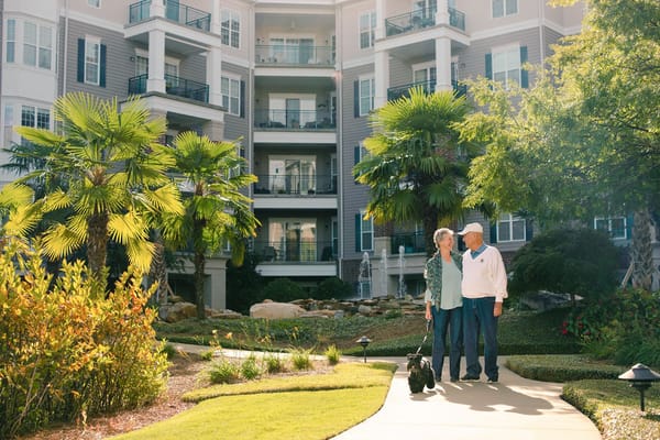 A senior couple walking a dog in a landscaped area near their residence.