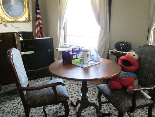 A round table with two chairs, one occupied by a plush Elmo toy, in a bright room with a window and decorative elements.