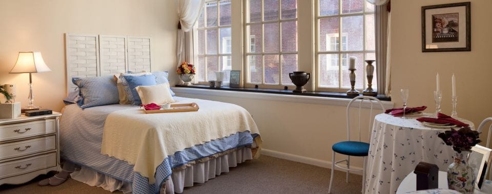 A well-furnished bedroom with a bed, nightstand, and dining table near a window.