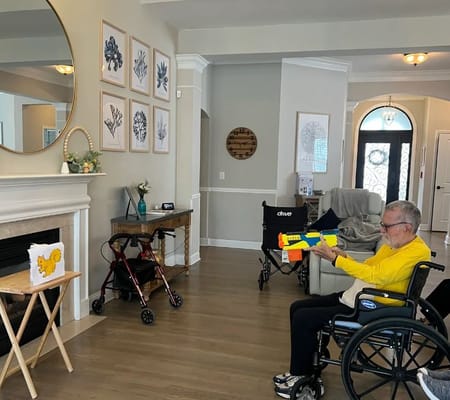 A smiling resident in a wheelchair holds a colorful toy gun in a bright living area.