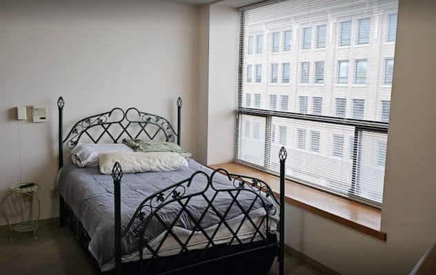 A neatly made bed in a small bedroom with large windows.