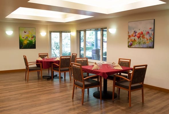 A dining room with red tablecloths and chairs, featuring large windows and artwork on the walls.