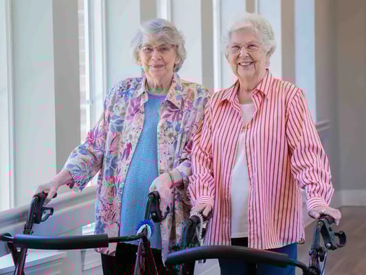 Two smiling residents using walkers in a bright hallway