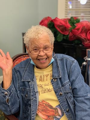 An elderly woman smiling and waving in a cozy setting