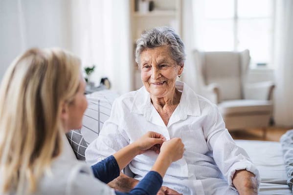 A caregiver assisting a smiling elderly woman indoors.