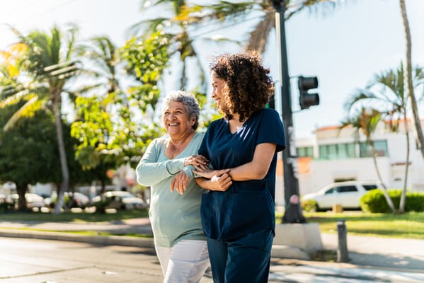 A senior woman walking with a caregiver in a sunny outdoor setting