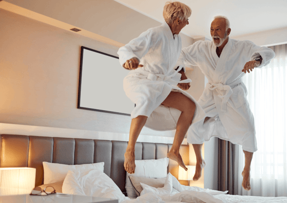 Two elderly people joyfully jumping on a bed