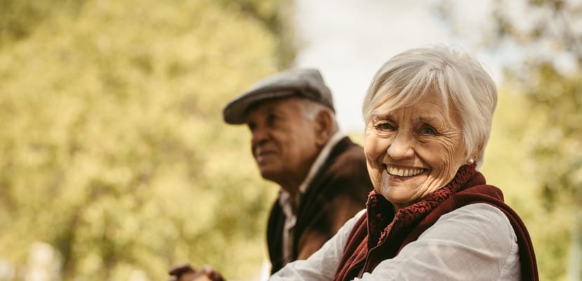 A smiling elderly woman with a man in the background outdoors