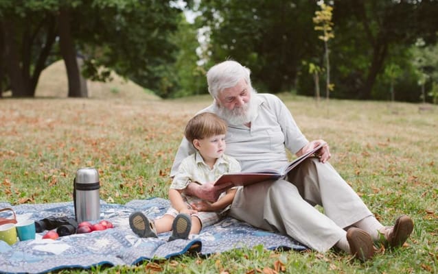 An elderly man reading to a young boy on a picnic blanket in a park