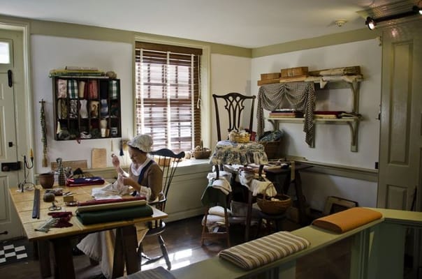 A woman sewing in a historical sewing room with various fabrics and tools.