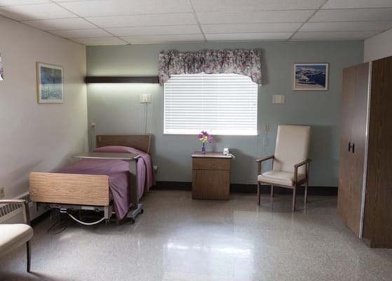 A bedroom with a bed, a chair, and a window in Franklin Plaza Extended Care.