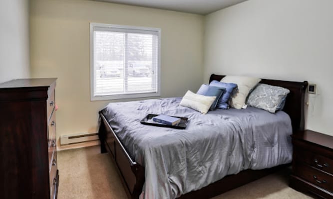 A cozy bedroom with a bed, dresser, and window at Minot Lodge Senior Living.