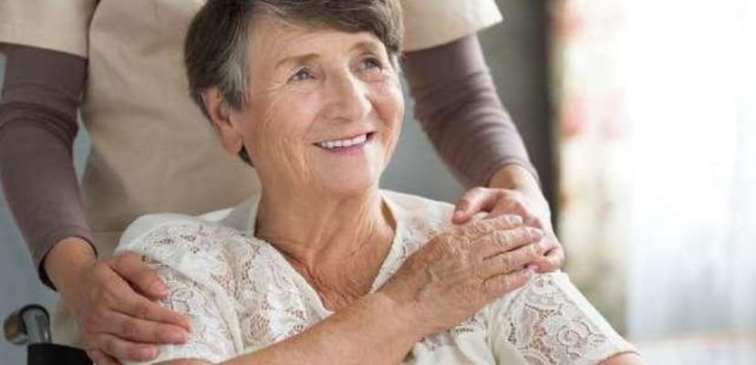 A smiling elderly woman in a wheelchair with staff assistance