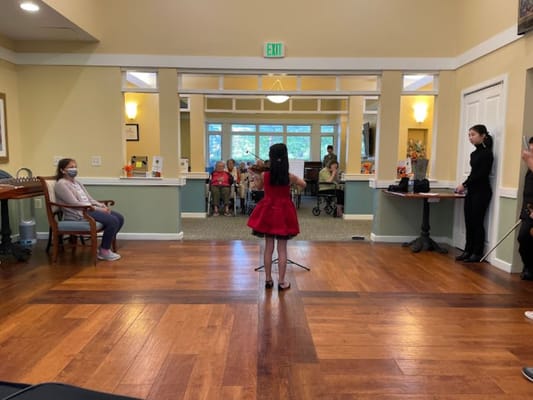 A young girl in a red dress performs with a violin in front of an audience at Raphael House.