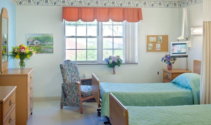 A well-lit bedroom with two beds, a floral chair, and a bouquet of flowers on a dresser.