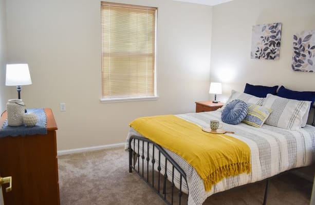 A cozy bedroom featuring a bed with a yellow throw and a bedside lamp.